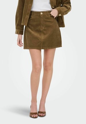 Femme portant une mini-jupe en velours côtelé marron, une veste assortie, un top blanc et des talons ouverts marron, debout devant un fond uni.