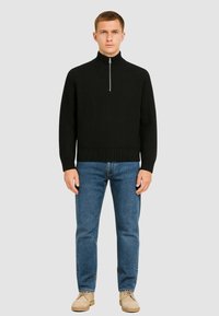 Maglione a coste nero con collo alto e zip argentata, abbinato a jeans in denim blu e mocassini beige. Modella in piedi su uno sfondo grigio.