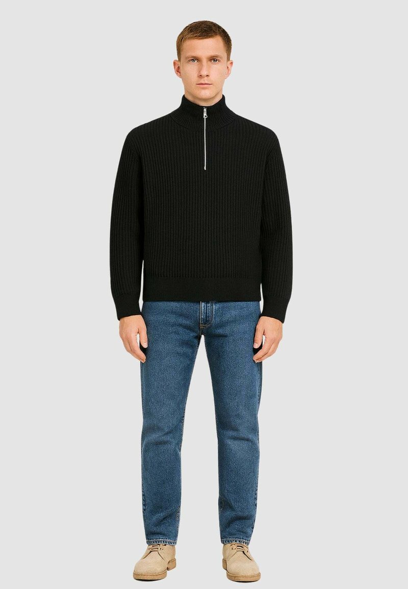 Maglione a coste nero con collo alto e zip argentata, abbinato a jeans in denim blu e mocassini beige. Modella in piedi su uno sfondo grigio.