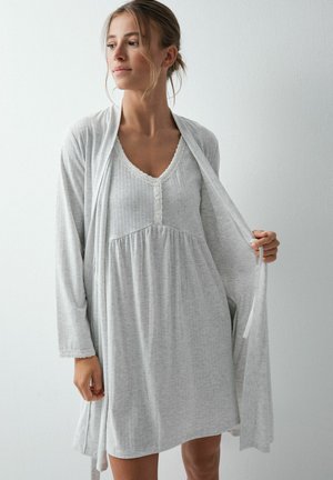 Femme portant une chemise de nuit gris clair à manches longues et une robe assortie, tenant la robe ouverte, debout contre un mur blanc.