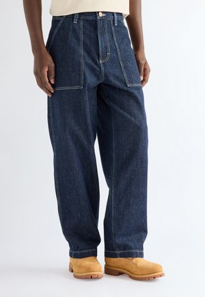 Jean denim bleu foncé à jambes larges porté avec des bottes de travail en daim beige sur une personne debout devant un fond blanc.