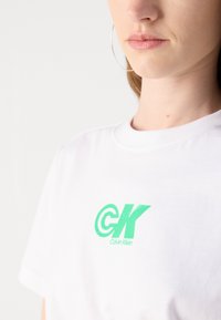 Balta medvilninė marškinėliai su dideliu žaliu "CK" logotipu ir "Calvin Klein" tekstu, su klasikiniu apvaliu kaklu ir švariu, minimalistiniu dizainu.