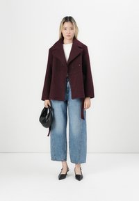Burgunderfarvet tekstureret jakke med dobbeltradet design, brede klapper og bånd i siden, parret med lysblå vide jeans og sorte accessories.