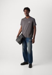 Grijze katoenen t-shirt met logo, blauwe denim jeans en zwarte schoenen. Een zachte zwarte tas wordt in de linkerhand gehouden, met een gebogen ontwerp.