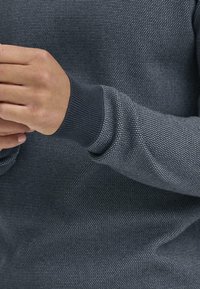 Sötét szürke kötött pulóver, texturált felülettel és bordázott mandzsettákkal. Feszes illeszkedésű, a szövet finom részleteivel.