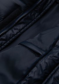 Tmavě modrá péřová bunda s texturovanou prošívanou tkaninou, která má hladké zapínání na zip a suchý zip jako detail uvnitř.