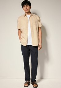 Camisa de manga corta beige con un patrón sutil, abotonada, combinada con pantalones azul marino oscuros y sandalias marrones. Camiseta blanca lisa debajo.
