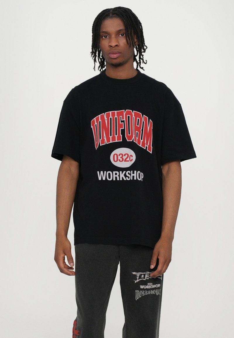Sort oversized t-shirt med rød og hvid grafisk tekst "UNIFORM 032c WORKSHOP." I bomuldsmateriale med en afslappet pasform.