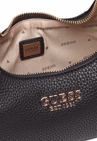 Intérieur en cuir texturé noir avec une fermeture éclair dorée. Présente une plaque logo GUESS et une doublure beige imprimée du motif 'GUESS'.