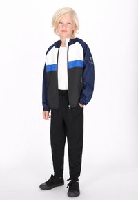 Blå og hvid farveblokeret letvægtsjakke med lynlåslukning, parret med sorte joggers og sorte sneakers. Glat stof, sporty design.