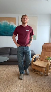 Homem em t-shirt e jeans maroon da Levi's, de pé sobre um tapete de palha na sala de estar, com sofá cinza e cadeira de vime.