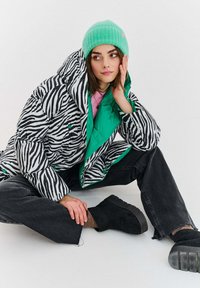 Prošívaná bunda se zebrovaným vzorem a zeleným vnitřkem, doplněná černými džínami a černými botami. Modelka má na sobě mentolovou čepici.