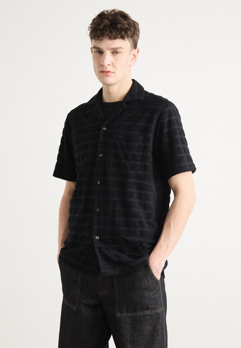 Svart t-skjorte med korte ermer, horisontale striper, strukturert stoff, knappelukking og brystlomme. Kombinert med mørke denimbukser.