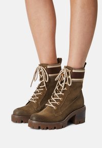 Braune Wildleder-Ankleboots mit einem stabilen Absatz, ausgestattet mit goldenen Beschlägen, beigen Schnürsenkeln und einem gestreiften Strickbund für zusätzliche Textur.