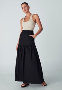 Gonna maxi nera con design arricciato e tasche, abbinata a una canotta beige a coste. Modella che indossa sandali neri, sfondo neutro.