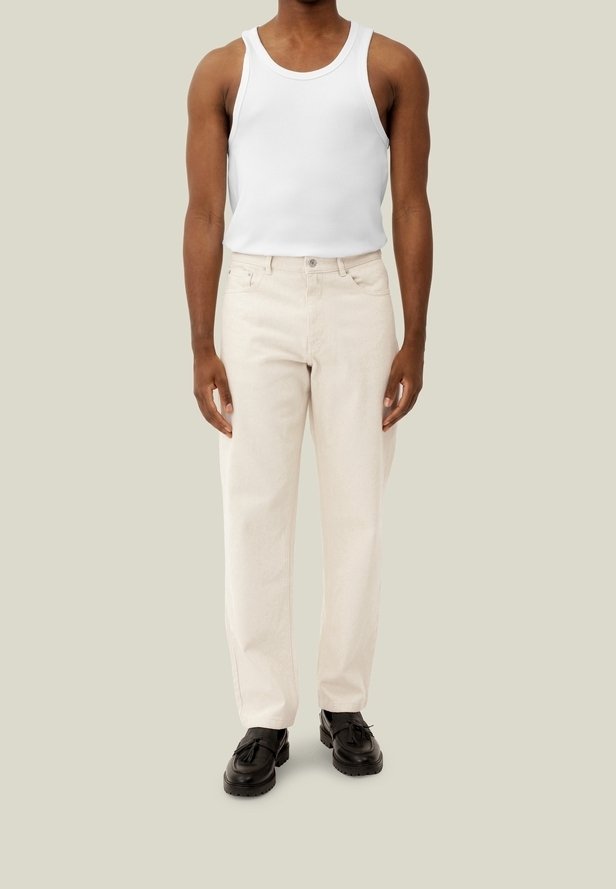 Jeans dritti color beige chiaro con fronte piatto, cinque tasche e chiusura con bottone metallico, abbinati a una canotta bianca e scarpe nere.