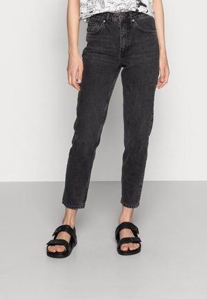 Person iført sorte højttaljede jeans og sorte åbne sandaler står på en grå gulv mod en hvid væg.