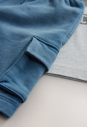 Kék pulóver mandzsetta, amely egy világosszürke, puha pamut anyagból készült ingre van helyezve. A pulóveren bordázott mandzsetta és varrási részletek találhatók.