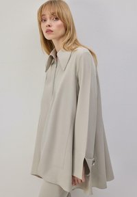 Blouse beige à manches longues avec col, dotée d'une fermeture éclair à l'avant, de fentes latérales et d'une coupe ample, fabriquée en tissu doux et lisse.
