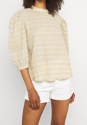 Vrouw draagt een beige gestructureerde blouse met pofmouwen en horizontale stiksels, gecombineerd met witte high-waisted shorts, staand tegen een effen achtergrond.