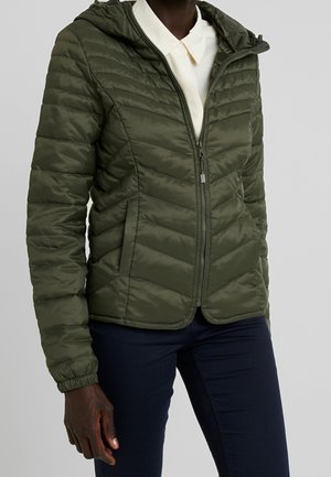 Osoba w oliwkowo-zielonej pikowanej kurtce z kapturem, zamkiem błyskawicznym, elastycznymi mankietami i bocznymi kieszeniami, ubrana na białą koszulę z kołnierzykiem i ciemne spodnie.