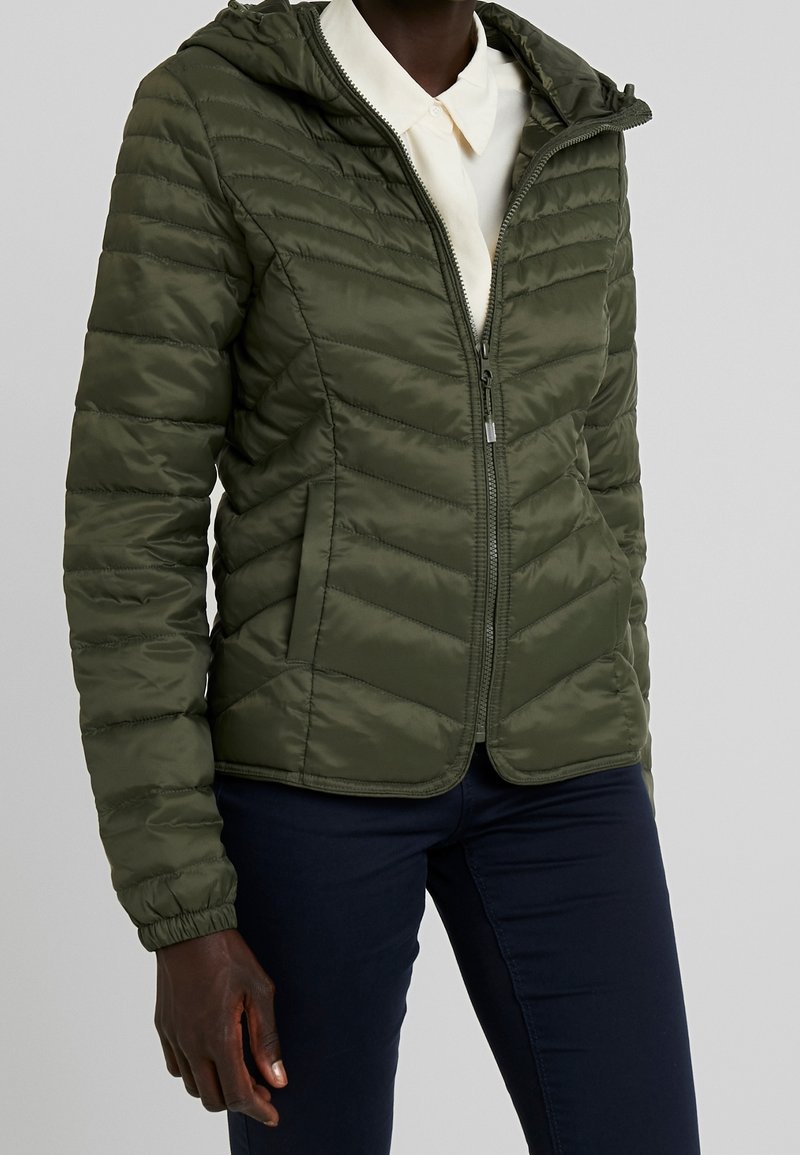 Personne portant une veste matelassée à capuche vert olive avec fermeture éclair, poignets élastiques et poches latérales, sur une chemise blanche à col et un pantalon foncé.