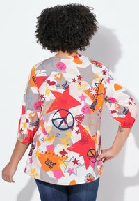 Farverig bluse med mønster, der viser stjerner, hjerter og fredssymboler i røde, orange og pink nuancer. Trekvart puffærmer, løs pasform.