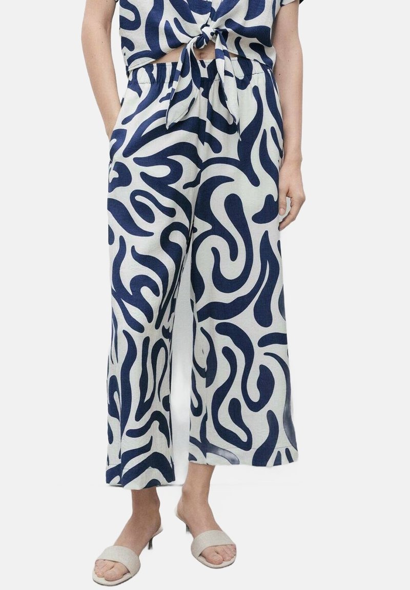 Béžové kalhoty culottes z látky s námořnickým abstraktním vzorem spirály. Obsahují elastický pas a široké nohavice. Vystavené s světle zbarvenými sandály.