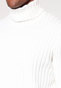 Vit ribbad polokrage-tröja gjord av tjockt stickat tyg. Har en hög krage och en figurnära silhuett med vertikal struktur.