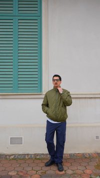 Hombre con chaqueta acolchada verde y pantalones azules de pie sobre un suelo con baldosas hexagonales, junto a una pared blanca con contraventanas azul verdoso.