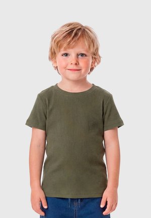 Uśmiechnięty młody chłopiec o blond włosach, ubrany w oliwkowozielony t-shirt i niebieskie dżinsy, stojący na tle jednolitego jasnoszarego tła.
