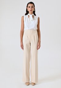 Witte mouwloze blouse met een geknoopt halsdetail, gecombineerd met hoge beige broeken. Gladde stof, geplooid ontwerp en slim fit.