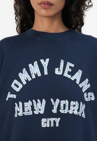 Felpa blu navy con collo a giro, con caratteri grandi in bianco e azzurro chiaro dal look consumato che recitano "TOMMY JEANS NEW YORK CITY."