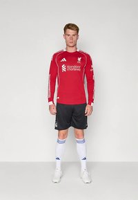 Equipamento de futebol vermelho de mangas compridas com o logótipo do Liverpool, combinado com calções pretos, meias brancas com riscas azuis e chuteiras brancas.