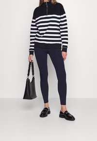 Sudadera con cremallera a rayas en blanco y negro, leggings negros, bolso negro y mocasines de charol. Tejido suave, corte relajado y texturas contrastantes.