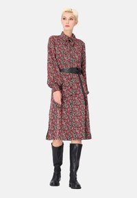 Robe florale rouge avec col, manches longues et ceinture. Le matériau est léger, avec des accents noirs et une coupe mi-longue. Associée à des bottes noires jusqu'aux genoux.