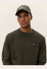 Olivgrön stickad tröja med ribbad halsringning, tillsammans med en ljusgrön keps med broderi av "Lower West Side".