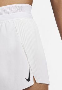 Baltos spalvos klostuotos sportinės kelnaitės su juodu Nike logotipu; turi tekstūruotą juosmenį ir šoninius plyšius geresniam judėjimui.