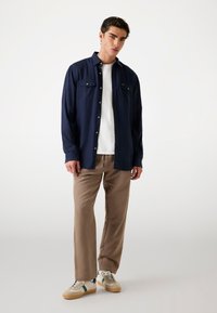 Eine marineblaue Hemdbluse mit langen Ärmeln, zwei Brusttaschen, kombiniert mit beigen Hosen und cremefarbenen Sneakers mit grünen Akzenten.