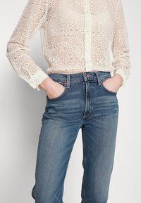 Camisa de botones de encaje en color blanco roto con intrincados patrones florales, combinada con unos pantalones vaqueros de mezclilla azul oscuro que cuentan con bolsillos frontales y costuras visibles.