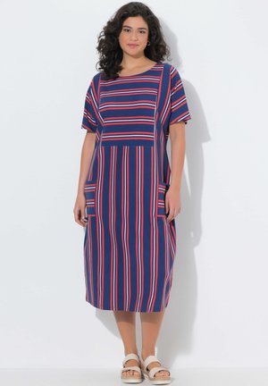 Kobieta ubrana w niebieską sukienkę midi z czerwonymi i białymi pionowymi oraz poziomymi paskami, krótkimi rękawami, dwoma przednimi kieszeniami i białymi sandałami.