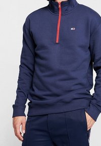 Marinblå fleecepullover med hög krage och halv dragkedja. Har en röd dragkedja och en liten logotyp på bröstet. Mjuk textur.