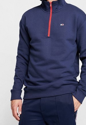 Navyblauwe fleece pullover met een hoge kraag en halve rits. Voorzien van een rode rits en een klein logo op de borst. Zachte textuur.