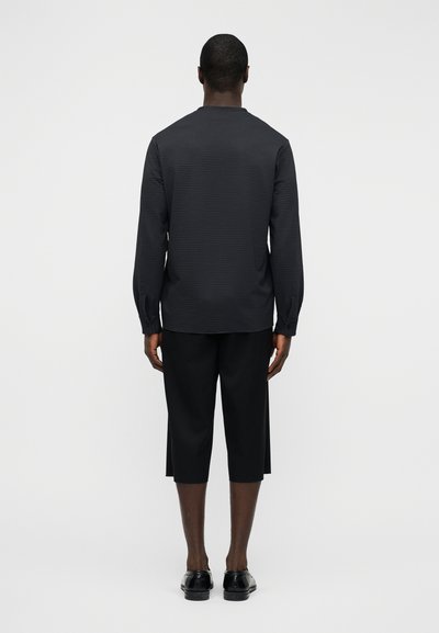 Μακρυμάνικο, υφασμάτινο σκούρο πουκάμισο με στρογγυλή λαιμόκοψη, συνδυασμένο με μαύρο παντελόνι μέχρι τα γόνατα και μαύρα παπούτσια, προβεβλημένο από πίσω.