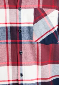Sección de camisa a cuadros roja, blanca y azul marino con cuatro botones negros y un bolsillo en el pecho en el lado derecho.