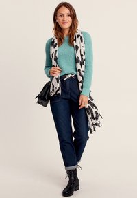 Un pull turquoise avec un tricot texturé, associé à un jean en denim foncé et des bottes noires à lacets. Une écharpe à motifs noir et blanc souligne la tenue.