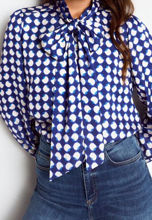 Kobieta nosząca niebieską bluzkę w białe i jasnoniebieskie kropki, związaną kokardą przy szyi, w połączeniu z niebieskimi jeansami z wysokim stanem.