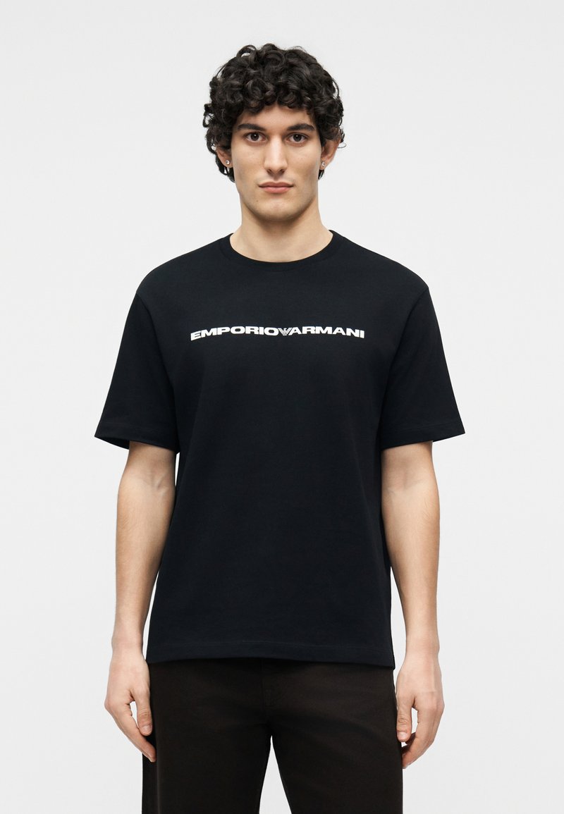 Νέος άνδρας με σγουρά μαλλιά που φοράει μαύρο t-shirt Emporio Armani με λευκό λογότυπο, στέκεται μπροστά σε απλό φωτεινό φόντο.