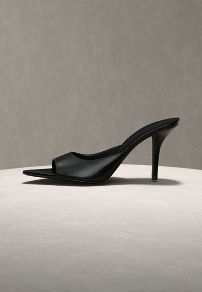 Μαύρο δερμάτινο mule με μυτερή μύτη, ανοιχτό μπροστινό μέρος και λεπτό ψηλό τακούνι, εμφανίζεται σε φωτεινή επιφάνεια με ουδέτερο φόντο.