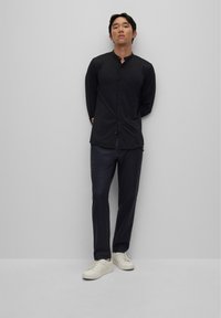 Zwarte longsleeve shirt met een staande kraag en knopen. Gecombineerd met donkere broek en witte sneakers, wat een strakke, minimalistische uitstraling benadrukt.